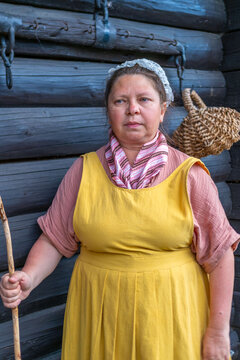 woman in traditional peasant costume, yellow pinafore dress, pink blouse, patterned scarf, lace headpiece, Historical portrait, holding wooden stick, near dark log cabin wall, basket, rural heritage