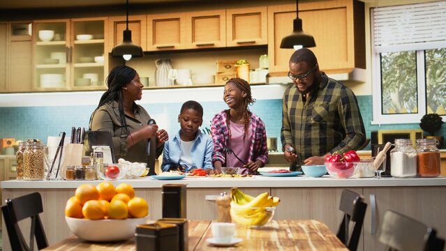 African American family enjoys a playful karaoke session during pizza day in the kitchen, singing with utensils and dancing together while cooking at home.