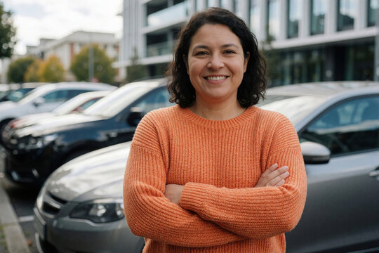 Smiling woman stands outdoors with arms crossed in New Zealand. She wears an orange sweater near parked cars with modern buildings in background. Confident woman happy lifestyle.