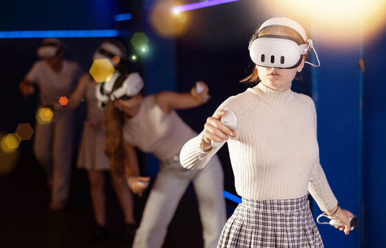 Excited young girl in VR glasses interacting with immersive environment of virtual reality world, standing with controllers in hands and actively gesticulating while controlling objects in game .