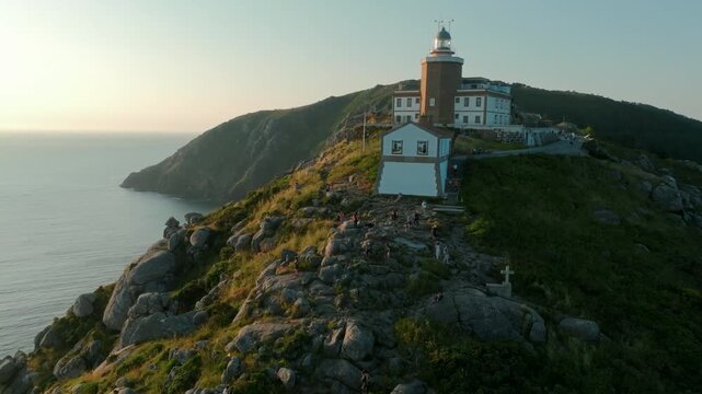 Serene aerial capture of the Cape Finisterre lighthouse at sunset, with pilgrims and travelers gathered on the rocky, windswept point, marking the emotional and geographic end of the Camino de