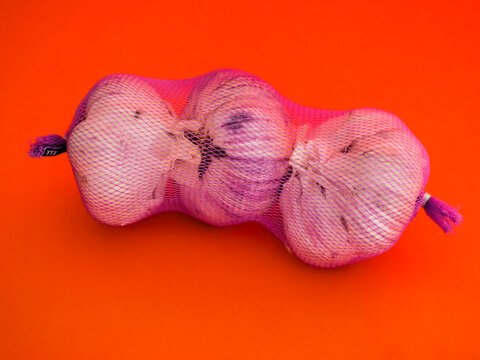 Three garlic bulbs rest in mesh bag, vibrant orange background. Bulbs show natural skin texture with purple tints. Bright lighting highlights garlic's form. Suitable for food, cooking