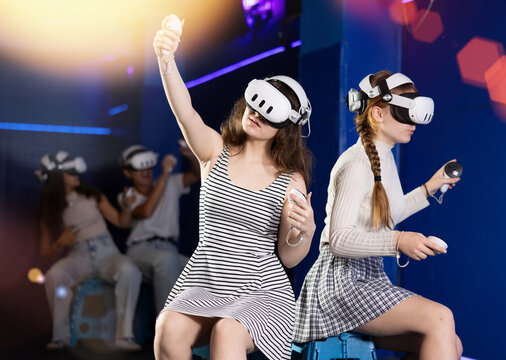 Cheerful young female friends wearing VR headsets and holding controllers having fun in virtual reality studio, competing in two-on-two shooter game