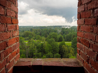 Brick window frames lush green forest under stormy sky. View invites quiet reflection or rustic charm. Ideal for travel, nature, or architectural themes. Mood: calm, contemplative, slightly moody © mark_gusev