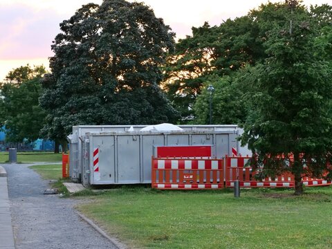 Industrial Waste Dumpster Containers Behind Red Safety Fences in Green City Park at Sunset, Construction and Park Maintenance Work Zone Concept
