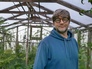 Man stands in wooden greenhouse with glass panels. He wears blue hoodie and glasses and he is in his 40s or 50s with grey beard and hair. Hands on hips, looks at camera. Growing organic produce. © mark_gusev
