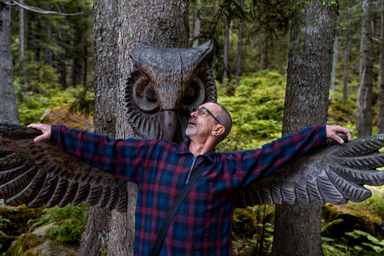 Man posing with outstretched arms mimicking a large wooden owl carving's wings in nature