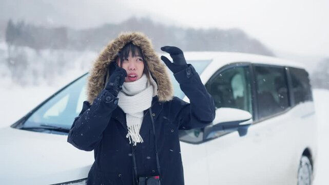 Asian woman calling roadside assistance or car insurance after vehicle breakdown in heavy snow during winter road trip travel countryside, stranded driver seeking emergency help in cold snowy weather