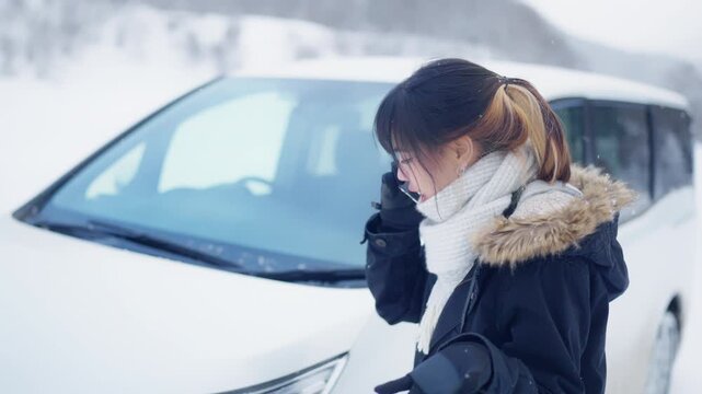 Asian woman calling roadside assistance or car insurance after vehicle breakdown in heavy snow during winter road trip travel countryside, stranded driver seeking emergency help in cold snowy weather