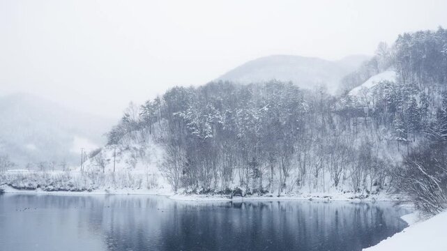 Snow covered lake with forest and mountain in winter, serene snowy landscape with calm water reflection and gentle snowfall in cold nature scenery