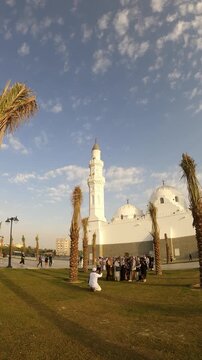 People gathering on the lawn outside the white Masjid Quba with palm trees under cloudy sky.
