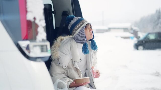 Asian man eating takeaway food in car trunk during winter road trip in snowy countryside. Male traveler enjoy outdoor active lifestyle taking meal break while traveling by vehicle on holiday vacation