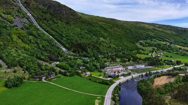 Aerial view of a hydroelectric power plant with a long penstock running down a lush green hillside next to a river and small village in Norway.
