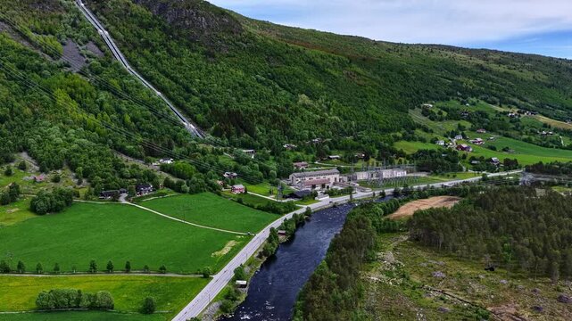 Aerial view of a hydroelectric power plant with a long penstock running down a steep green mountain slope, a river, and a rural road in Norway.
