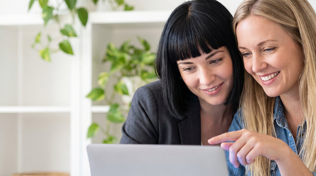 Two diverse female colleagues smiling and working together on laptop computer in bright office, businesswomen pointing at screen, collaboration and teamwork concept