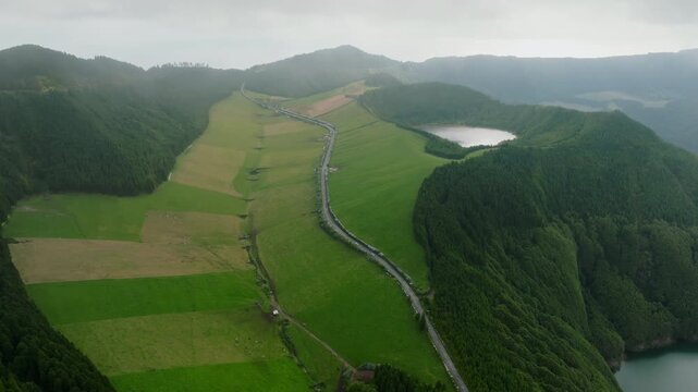 Aerial view of a winding road traversing lush green volcanic pastures and agricultural fields, adjacent to a smaller crater lake and dense forest slopes within the Sete Cidades caldera, S o Miguel