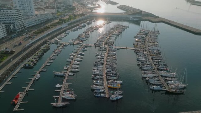 Aerial view of the Ponta Delgada marina at sunrise, showcasing a densely packed harbor of sailboats and yachts, reflecting the golden light, adjacent to the city's waterfront and urban architecture, S