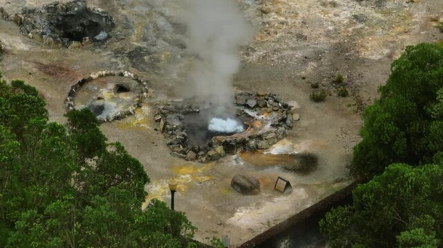 Aerial close-up of active geothermal fumaroles in Furnas, S o Miguel Island, showing steaming volcanic vents surrounded by mineral-rich rocky ground and lush green vegetation.