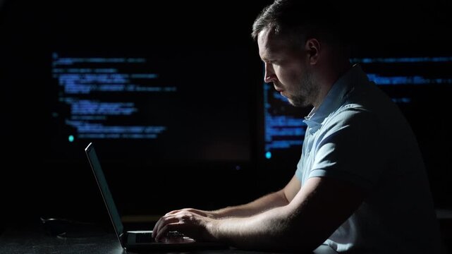 Programmer types code on laptop in darkness. Programmer works beside illuminated computer screens. Blue code screens light programmer from behind. Side view frames programmer using modern laptop.