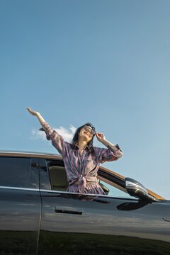 A woman leans out of a car window, smiling and raising her hands in the air while surrounded by clear blue skies. The setting is an open countryside landscape, highlighting a carefree day outdoors
