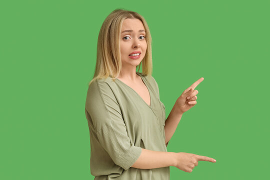 Young woman feeling embarrassment while pointing at something on green background