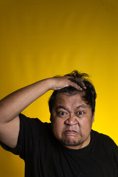 An intensely frustrated young Asian male aggressively grabs the hair with one hand while presenting a fiercely scrunched facial expression against a solid yellow background. The angry