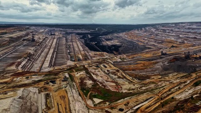Aerial view of the vast industrial landscape of the Tagebau Hambach open-pit coal mine with deep excavation terraces and heavy machinery under a cloudy sky in Elsdorf, Nordrhein-Westfalen, Germany.