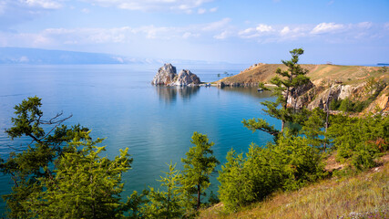 Obraz premium Wide landscape view of a blue Lake Baikal and prominent two-peaked rock formation. Green coniferous trees on a grassy cliff coast. Summer travel destination in nature