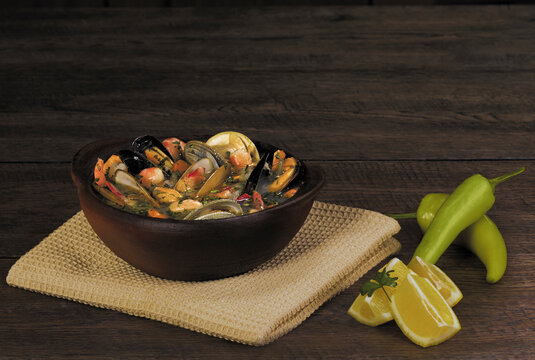 Chilean mariscal seafood soup served in a traditional clay bowl (paila de greda) with mussels, clams, and shrimp, accompanied by lemon wedges and green chili pepper on a dark wooden table 