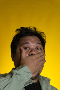 A striking studio portrait features a surprised young Asian male covering a mouth with one hand against a vibrant yellow background. The individual gazes directly into the lens with