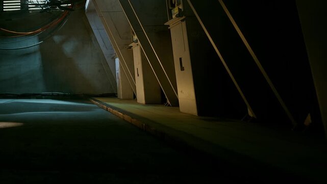 A hallway is shown with dim lighting and shadows. Utility boxes are placed along the wall. The setting appears to be an underground area. There are wires dangling from the ceiling.
