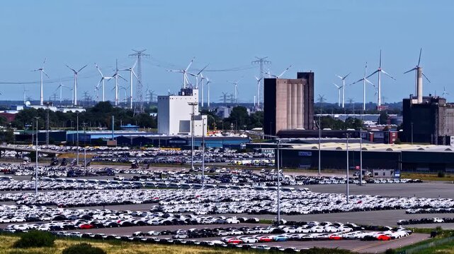Aerial view of the Volkswagenwerk Emden factory with rows of new cars in the parking lot and wind turbines in the background in Emden, Niedersachsen, Germany.