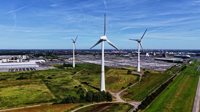 Aerial view of wind turbines near the Volkswagenwerk Emden industrial facility with vast car storage lots and green fields under a clear blue sky in Emden, Niedersachsen, Germany.