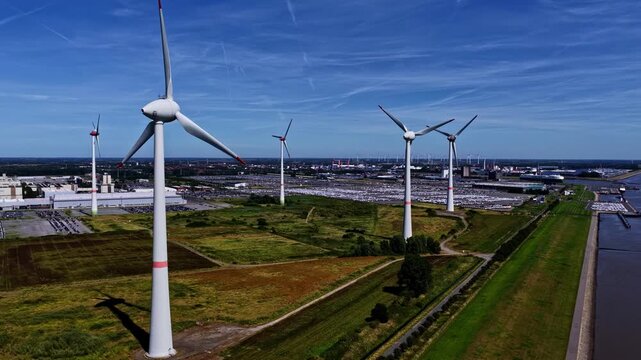 Aerial view of wind turbines at the Volkswagenwerk Emden factory with large parking lots filled with cars and a river nearby in Emden, Niedersachsen, Germany.