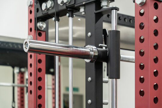 Close-up view of a barbell resting on a smith machine in a fitness gym, highlighting the metal bar, safety supports, and red and black steel frame.