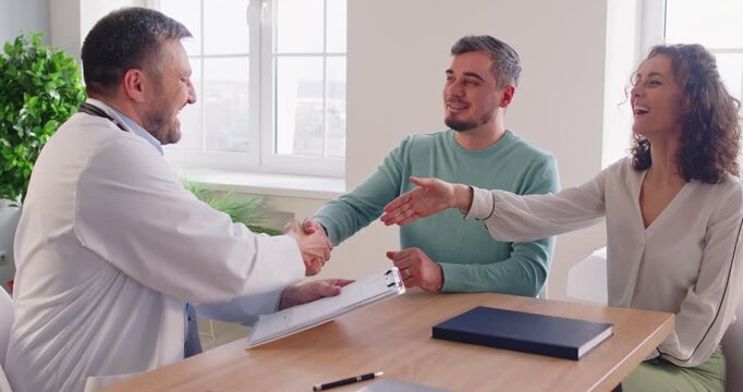 Smiling doctor shaking hands with cheerful couple planning pregnancy in medical office. Friendly healthcare professional talking about successful IVF treatment, congratulating young happy family.