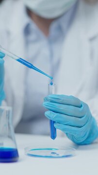 Close-up of a laboratory technician using a pipette to transfer blue liquid sample into a test tube in a clean scientific laboratory environment.