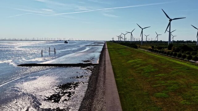Aerial view of wind turbines along the coastal dike and the Ems estuary with calm water and green grass in Emden, Niedersachsen, Germany.