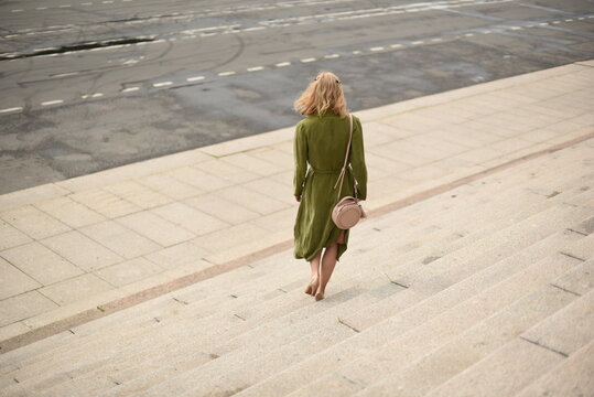 Rear view of stylish blonde woman in green coat walking down wide stone stairs in city