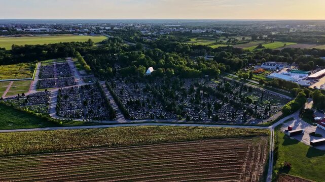 Aerial view of a cemetery with rows of graves, surrounded by green trees and agricultural fields in Plock, Mazowieckie, Poland.