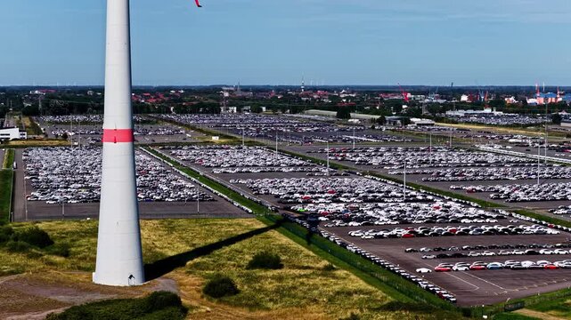 Aerial view of the Volkswagenwerk Emden car storage lot filled with rows of new vehicles next to a large wind turbine in Emden, Niedersachsen, Germany.