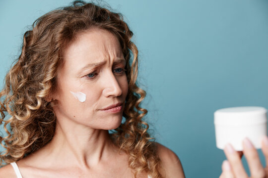Authentic beauty of a middle-aged woman with natural skin without retouching. She examines cream jar with doubt. Concept for skincare, aging, and realistic beauty care.