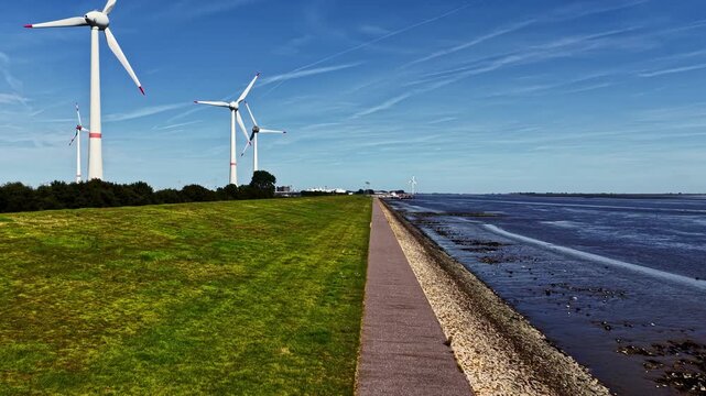 Aerial view of wind turbines along the Ems river with a paved path and green grass in Emden, Niedersachsen, Germany.