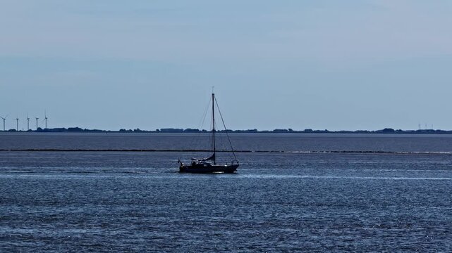 Aerial view of a lone sailboat gliding across the calm water with wind turbines visible on the distant horizon in Emden, Niedersachsen, Germany.