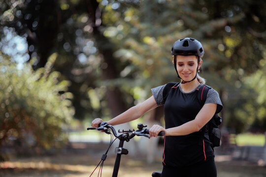 Happy Female Cyclist In Helmet Looking Down At Her Electric Bicycle. Smiling young woman adjusts her e-bike on a path in a green park