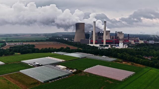 Aerial view of RWE Power AG power plant Niederaussem with cooling towers emitting steam and large greenhouses in the foreground in Bergheim, Nordrhein-Westfalen, Germany.