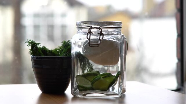 Person placing fresh cucumber slices into glass pickling jar with dill and garlic indoors.