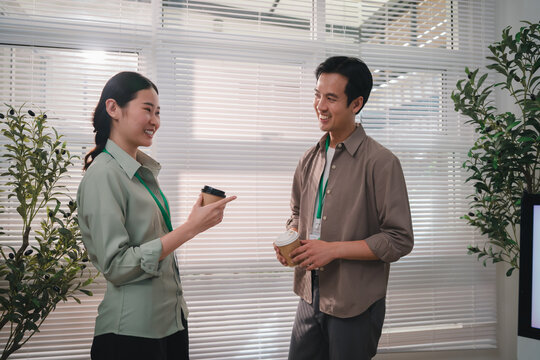 esg concept, Asian coworkers discussing work in bright office while holding smartphone and takeaway coffee with relaxed expressions