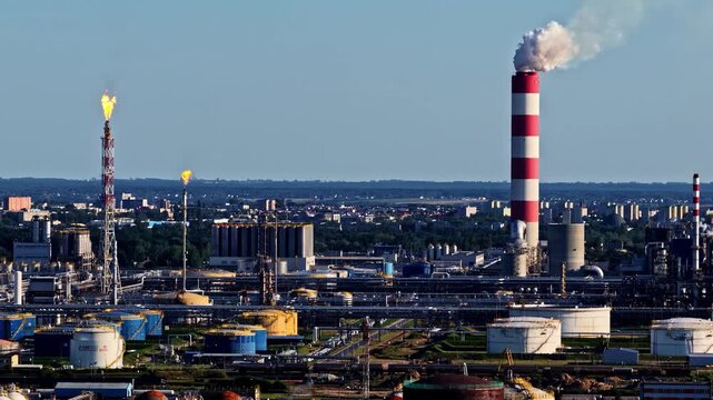 Aerial view of the Orlen Plock Production Plant with industrial chimneys, storage tanks, and flares emitting smoke against a clear blue sky in Plock, Mazowieckie, Poland.