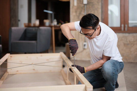 Asian mature man concentrates on self-assembling wooden furniture with DIY tools, tightening strength with a screwdriver, home improvement, hobby carpentry workshop, expert handyman, active lifestyle.